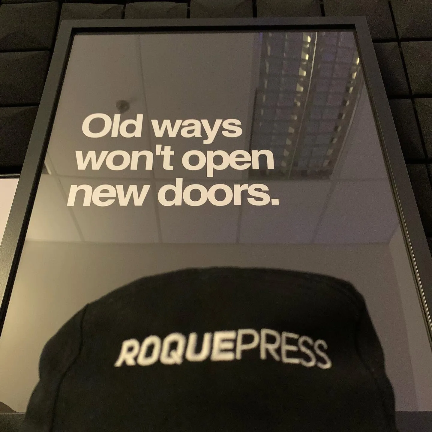 Framed print reading “Old ways won't open new doors.” above a black Roquepress cap